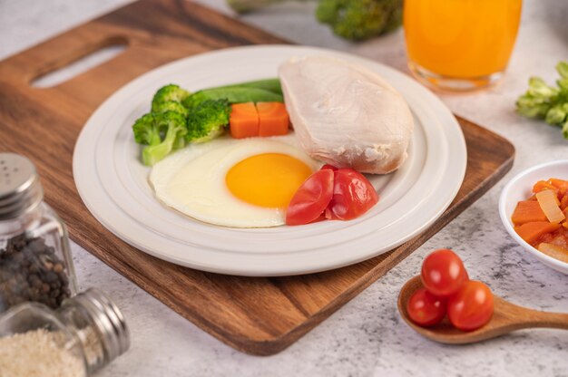 Breakfast consists of chicken, fried eggs, broccoli, carrots, tomatoes and lettuce on a white plate.