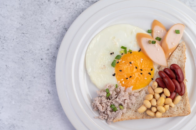 Breakfast consists of fried eggs, sausage, minced pork, bread, red beans and soy on a white plate.