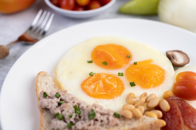 朝食は目玉焼き、ソーセージ、豚ひき肉、パン、小豆、大豆の白い皿で構成されています。