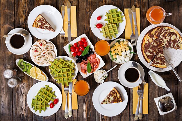 Breakfast food table. Festive brunch set, meal variety with spinach waffles, salmon, cheese, olives, chicken rolls and cheesecake. Top view. Flat lay