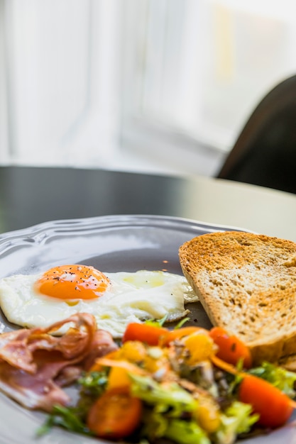 Breakfast gray plate with egg; bacon; toast and salad on table