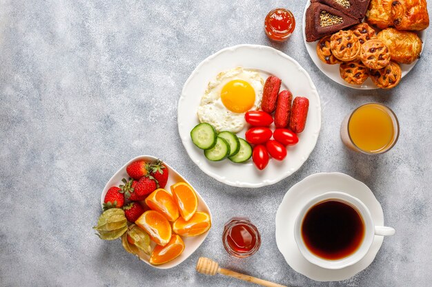 A breakfast plate containing cocktail sausages,fried eggs,cherry tomatoes,sweets,fruits and a glass of peach juice.