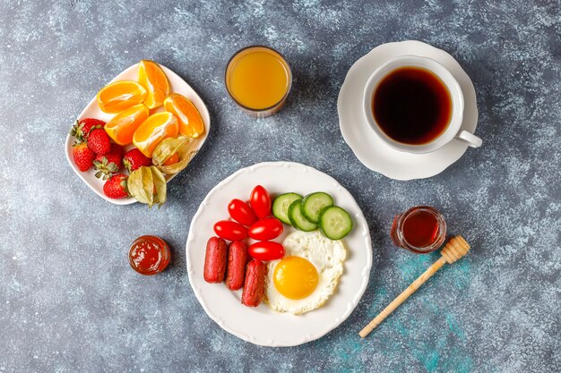 A breakfast plate containing cocktail sausages,fried eggs,cherry tomatoes,sweets,fruits and a glass of peach juice.