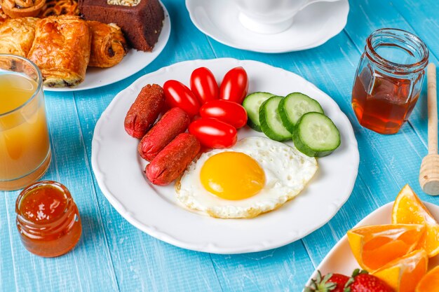 A breakfast plate containing cocktail sausages,fried eggs,cherry tomatoes,sweets,fruits and a glass of peach juice.