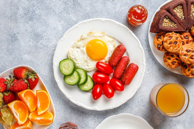 A breakfast plate containing cocktail sausages,fried eggs,cherry tomatoes,sweets,fruits and a glass of peach juice.