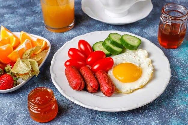 A breakfast plate containing cocktail sausages,fried eggs,cherry tomatoes,sweets,fruits and a glass of peach juice.