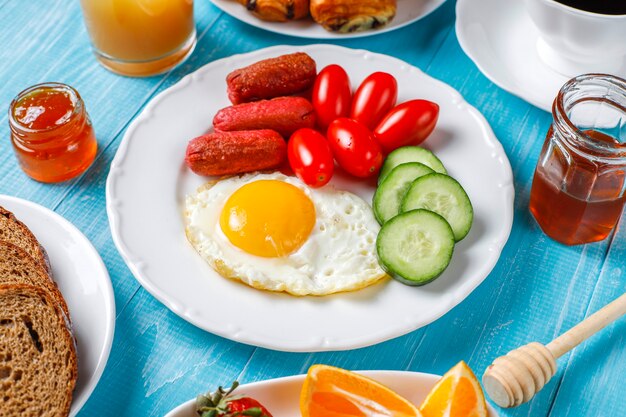 A breakfast plate containing cocktail sausages,fried eggs,cherry tomatoes,sweets,fruits and a glass of peach juice.