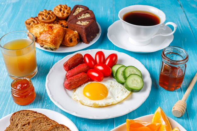 A breakfast plate containing cocktail sausages,fried eggs,cherry tomatoes,sweets,fruits and a glass of peach juice.