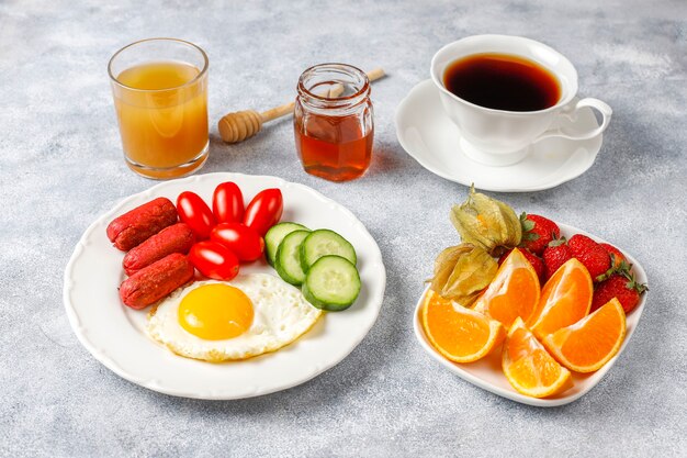 A breakfast plate containing cocktail sausages,fried eggs,cherry tomatoes,sweets,fruits and a glass of peach juice.