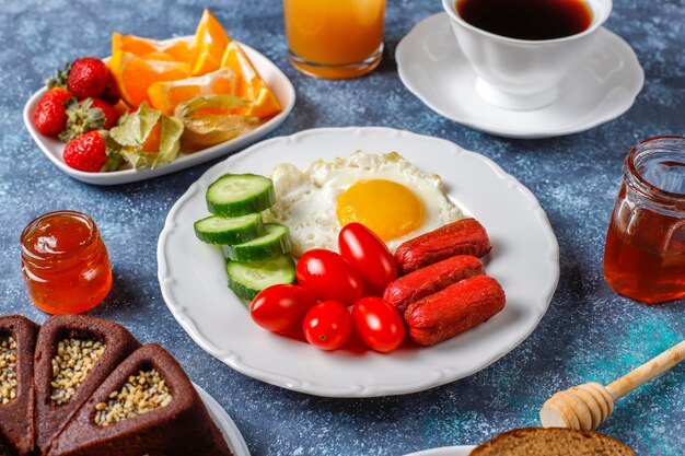 A breakfast plate containing cocktail sausages,fried eggs,cherry tomatoes,sweets,fruits and a glass of peach juice.