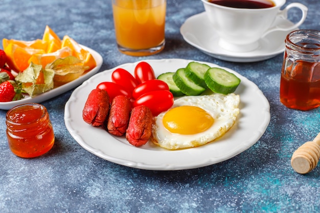 A breakfast plate containing cocktail sausages,fried eggs,cherry tomatoes,sweets,fruits and a glass of peach juice.