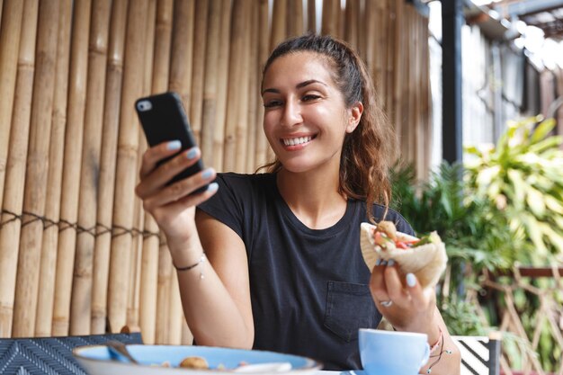 Breakfast in restaurant timemanagement and lifestyle concept Young female tourist travel blogger reading feedback under her new post internet hold mobile phone and pita eating at cafe