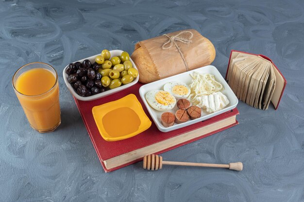 Breakfast set bundled on top of a book, next to a small notebook, a honey spoon and a glass of juice on marble surface.