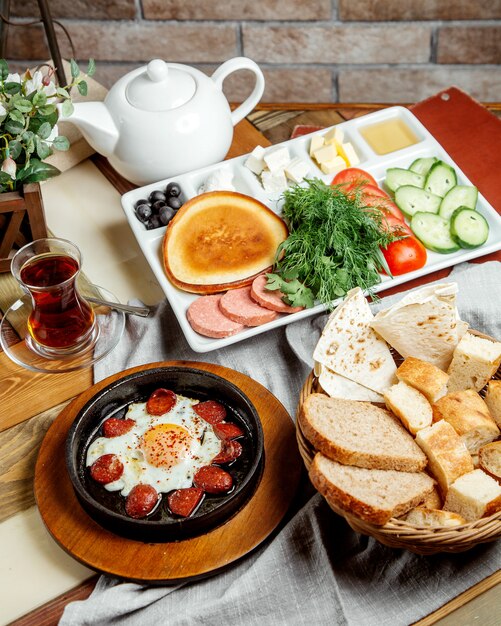 Breakfast setup with egg and sausage dish breads vegetable slices cheese honey and tea