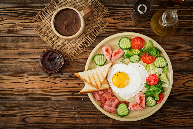 Breakfast on Valentine's Day - fried egg in the shape of a heart, toasts, sausage, baconnd fresh vegetables. English breakfast. Cup of coffee. Top view