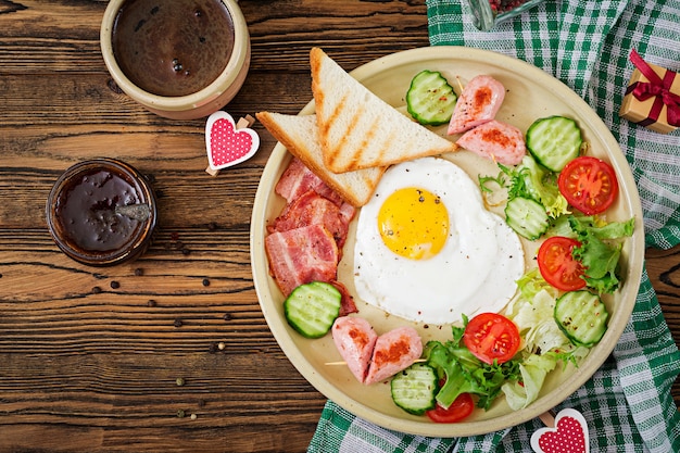 Breakfast on Valentine's Day - fried egg in the shape of a heart, toasts, sausage, baconnd fresh vegetables. English breakfast. Cup of coffee. Top view