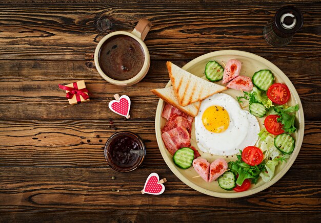 Завтрак на День святого Валентина - жареное яйцо в форме сердца, тосты, колбаса, бекон и свежие овощи. Английский завтрак. Чашка кофе. Вид сверху