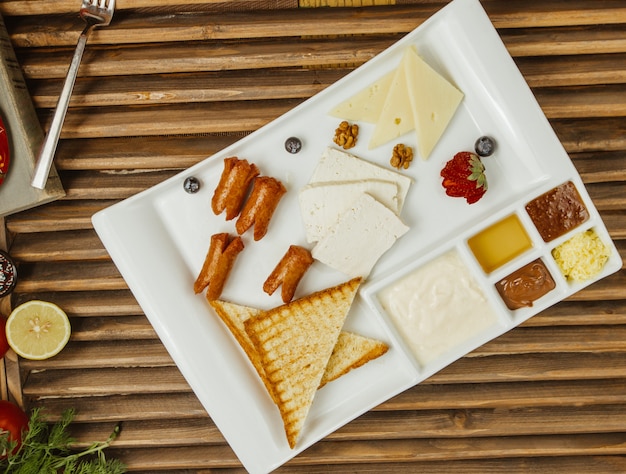 Деревянная доска для завтрака с блинчиками, медом, сливочным сыром, овощами и конфитюром в квадратной белой тарелке