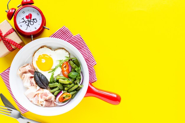 Breakfast for your beloved for the holiday: heart-shaped egg, bacon, green beans on a yellow background. Selective focus. View from above. Copy space.