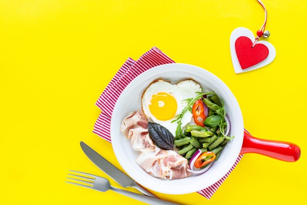 Breakfast for your beloved for the holiday: heart-shaped egg, bacon, green beans on a yellow background. Selective focus. View from above. Copy space