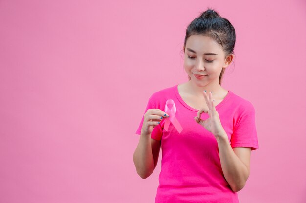 유방암 인식, 분홍색 셔츠를 입은 여성, 오른손으로 분홍색 리본을 들고 왼쪽 손은 유방암에 대한 일일 상징을 보여 주면서 괜찮습니다.