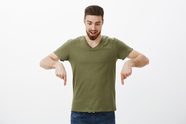 Breathtaking product right here. Portrait of amused and stunned impressed good-looking bearded male pointing and looking down with dropped jaw excited of seeing cool sale over white wall