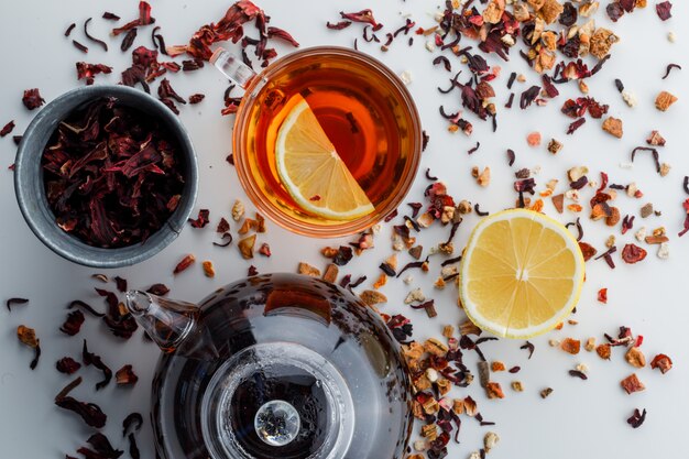 Brewed tea with dried herbs, lemon in glass and teapot on white surface