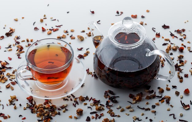 Заваренный чай со смешанными сушеными травами в чайнике и чашке на белой поверхности