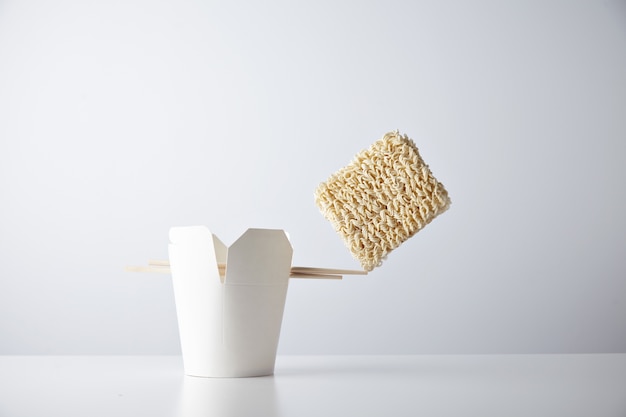 Free photo brick of dry noodles balances on the edge of chopsticks on blank takeaway box isolated on white commercial retail set