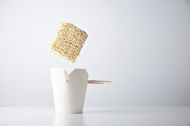 Brick of dry noodles falls down inside blank takeaway box with chopsticks isolated on white commercial retail set