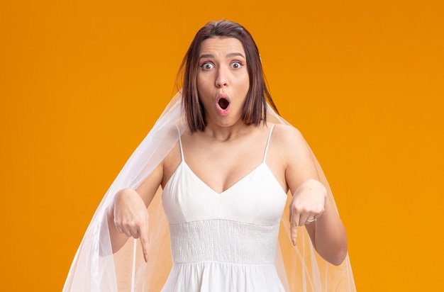 Bride in beautiful wedding dress amazed and surprised pointing with index finger down
