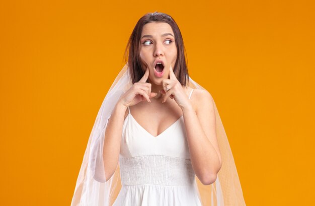 Bride in beautiful wedding dress looking aside being surprised pointing with index fingers at cheeks standing on orange