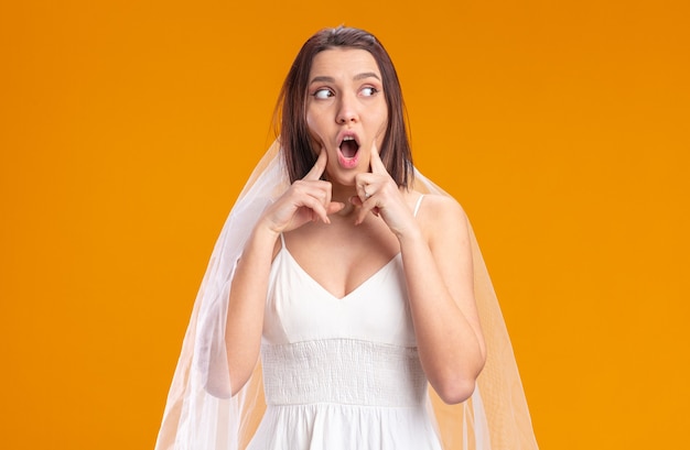 Bride in beautiful wedding dress looking aside being surprised pointing with index fingers at cheeks standing on orange