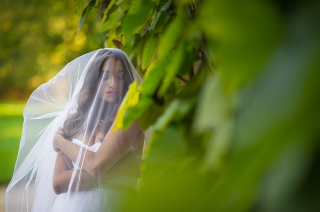 "Bride covered with veil standing at greenery"