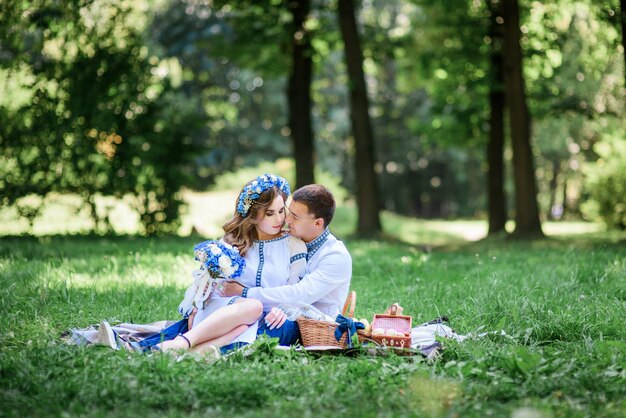 Bride and groom dressed in blue Ukrainian national clothes sit on the blanket 