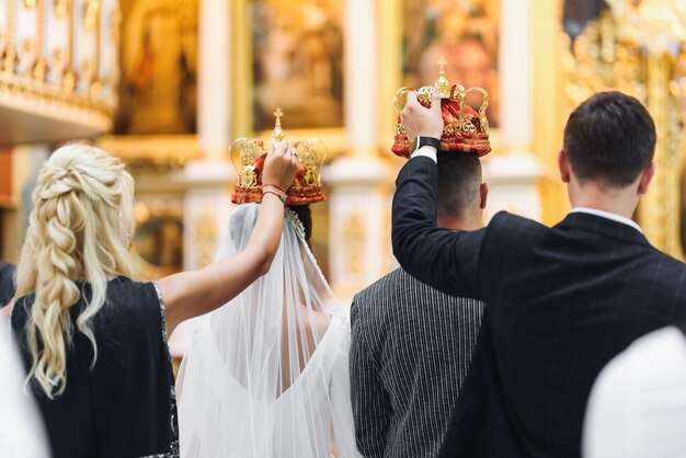 Жених и невеста стоят с коронами во время церемонии в церкви