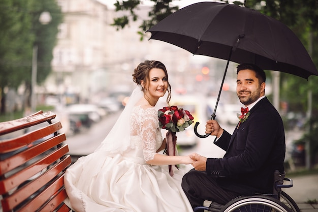 Bride and groom on the wheelchair sit kissing on the bench in the park