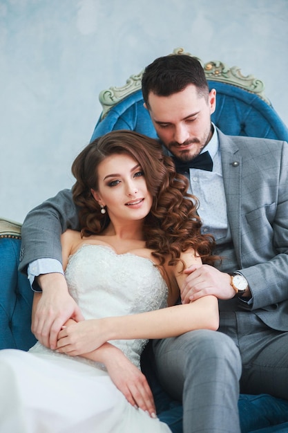 Невеста в красивом платье и жених в сером костюме сидят на диване в помещении в белом интерьере студии, как дома Premium Фотографии