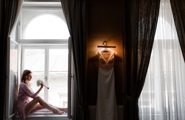 Bride is dressed in silky nightwear is sitting near window and holding wedding bouquet and wedding dress is hanging on the lamp