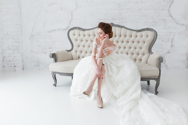 Bride in long dress sitting on sofa indoors in white studio interior like at home.