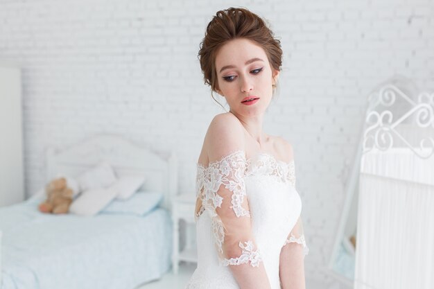 Невеста позирует в свадебном платье в студии на фоне кирпичной стены и белой кровати