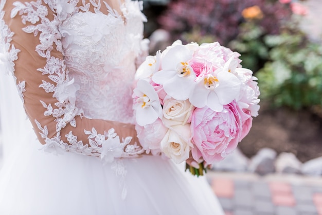 リッチドレスの花嫁は、蘭と牡丹のピンクの結婚式の花束を保持しています