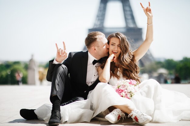 La sposa si siede a ridere prima della torre eiffel a parigi mentre lo sposo la bacia