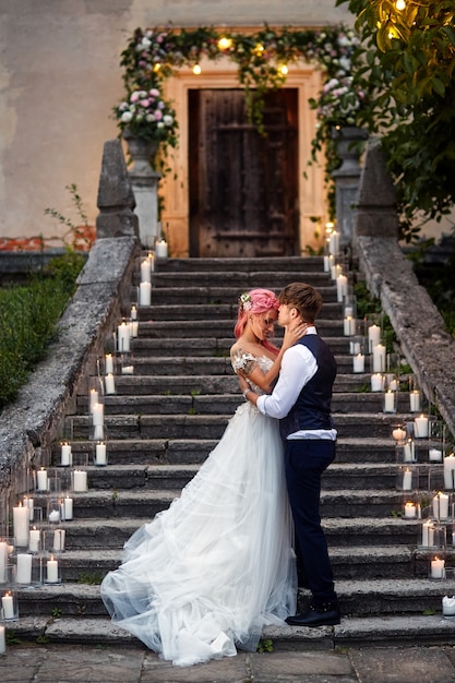 Bride with pink hair and stylish groom stand on footsteps with shiny candles