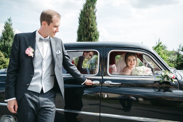 The bridegroom is standing by the car while the bride is in the car