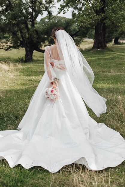 Brides stands in a park and holds a wedding bouquet behind her back, without face