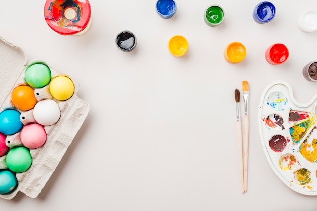 Bright collection of colored eggs near container near brushes, water colors and palette