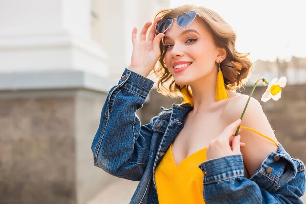 Яркий красочный портрет красивой молодой женщины, выглядящей в веселом настроении со счастливой улыбкой, в стильных хипстерских солнцезащитных очках, весенне-летней модной тенденции, джинсовой куртке, желтом топе