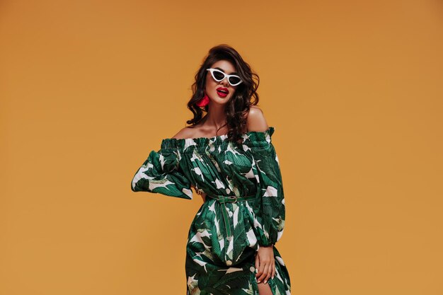 Bright curly girl with red lips and stylish earrings in cool sunglasses and green modern sundress on orange isolated background