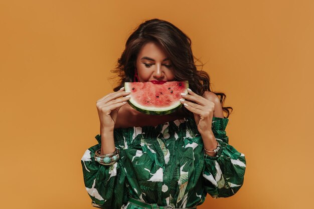 Bright curly haired lady with red lips in accessories and green fashionable clothes eating watermelon on isolated background